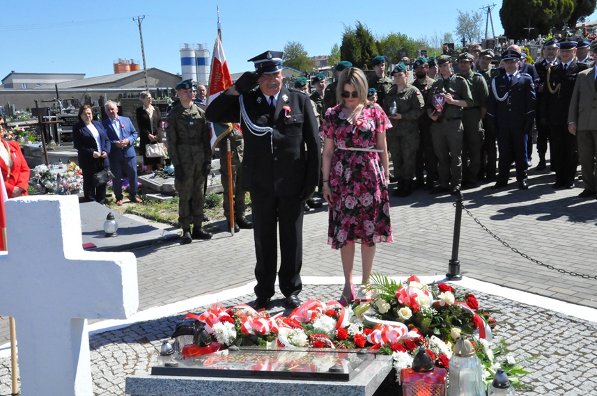Kobieta w sukience w różowe kwiaty i mężczyzna w mundurze strażackim składają hołd przed pomnikiem. Za nimi stoją osoby w mundurach i odświętnych strojach. Dalej nagrobki i kilka drzew. Pogodny dzień.