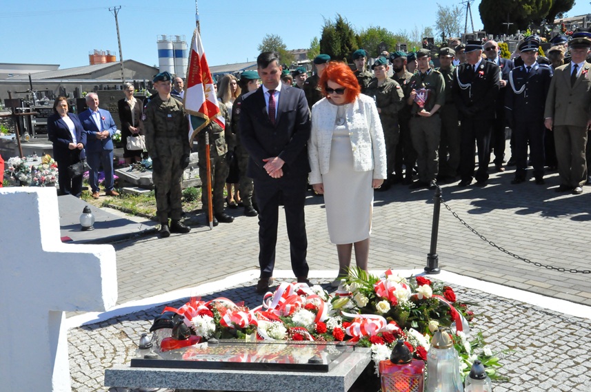 Kobieta w szarym kostiumie i mężczyzna w czarnym garniturze składają hołd przed pomnikiem. Za nimi stoją osoby w mundurach i odświętnych strojach. Dalej nagrobki i kilka drzew. Pogodny dzień.