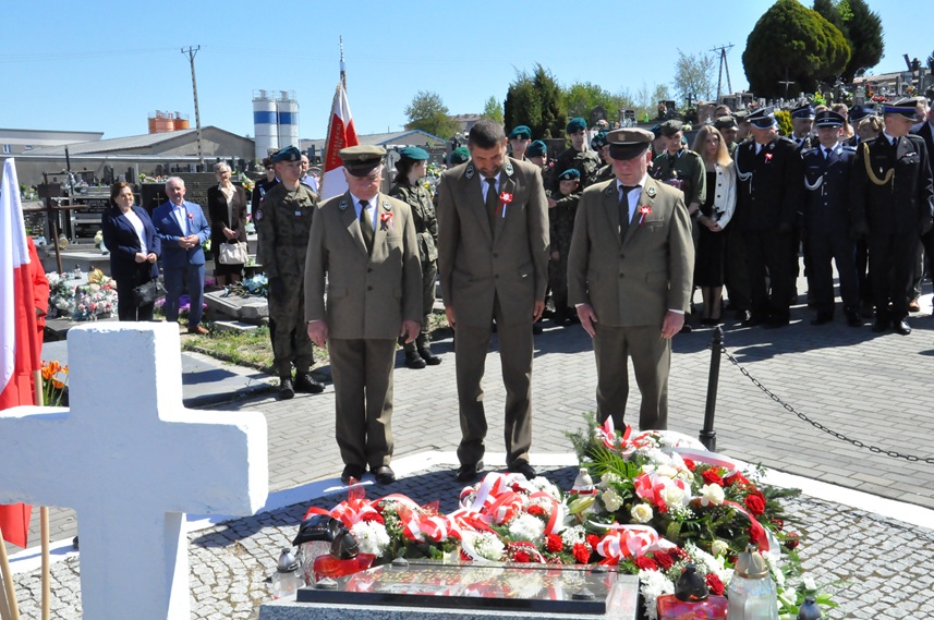 Trzech mężczyzn w mundurach leśników składa hołd przed pomnikiem. Za nimi stoją osoby w mundurach i odświętnych strojach. Dalej nagrobki i kilka drzew. Pogodny dzień.