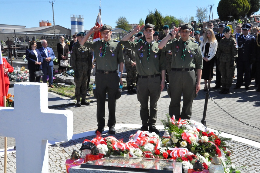 Trzech nastolatów w harcerskich strojach składa hołd przed pomnikiem. Za nimi stoją osoby w mundurach i odświętnych strojach. Dalej nagrobki i kilka drzew. Pogodny dzień.