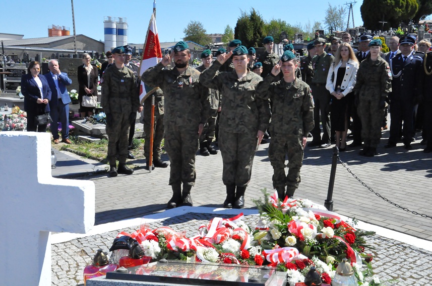 Trzy osoby w strzeleckich mundurach składa hołd przed pomnikiem. Za nimi stoją osoby w mundurach i odświętnych strojach. Dalej nagrobki i kilka drzew. Pogodny dzień.