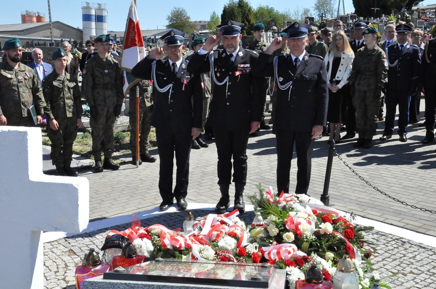 Trzech mężczyzn w strażackich mundurach składa hołd przed pomnikiem. Za nimi stoją osoby w mundurach i odświętnych strojach. Dalej nagrobki i kilka drzew. Pogodny dzień.