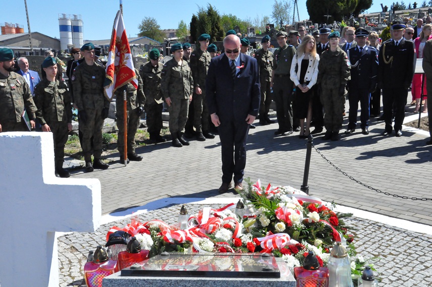 Mężczyzna w czarnym garniturze składa hołd przed pomnikiem. Za nimi stoją osoby w mundurach i odświętnych strojach. Dalej nagrobki i kilka drzew. Pogodny dzień.