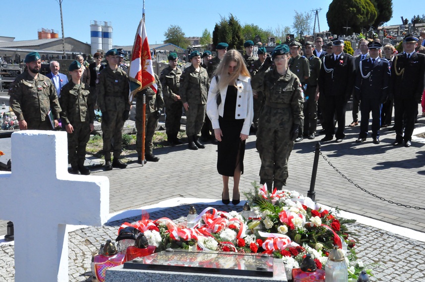 Dziewczyna w białej marynarce i dziewczyna w strzeleckim stroju składają hołd przed pomnikiem. Za nimi stoją osoby w mundurach i odświętnych strojach. Dalej nagrobki i kilka drzew. Pogodny dzień.