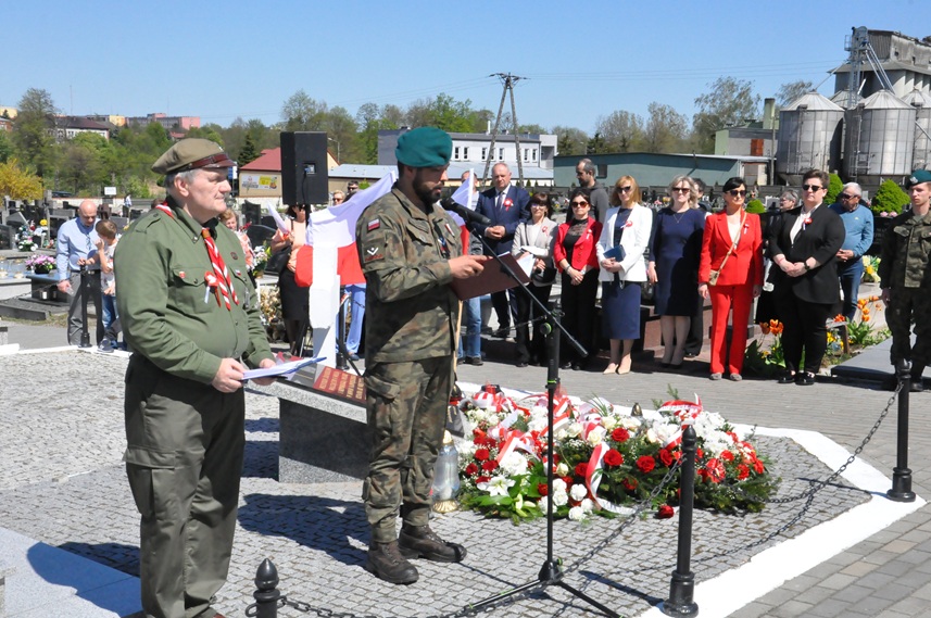 Mężczyzna w strzeleckim mundurze stoi przed mikrofonem na statywie. Obok niego stoi mężczyzna w mundurze harcerskim. Za nimi stoi więcej osób w odświętnych strojach. Dalej zabudowa miejska. Pogodny dzień.