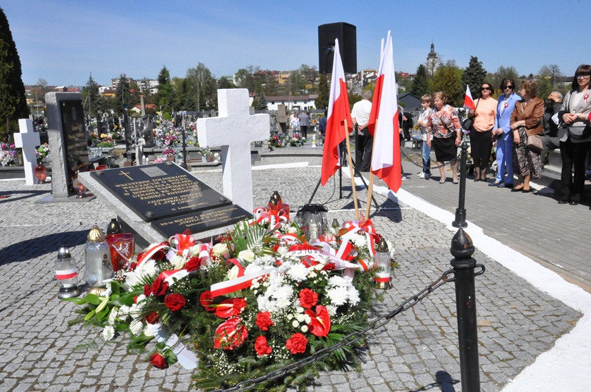 Wiązanki kwiatów w narodowych barwach i znicze przy pomniku. Obok stoi statyw z kilkoma flagami Polski. W dalszym planie kilka osób w odświętnych strojach, nagrobki oraz panorama miasteczka.