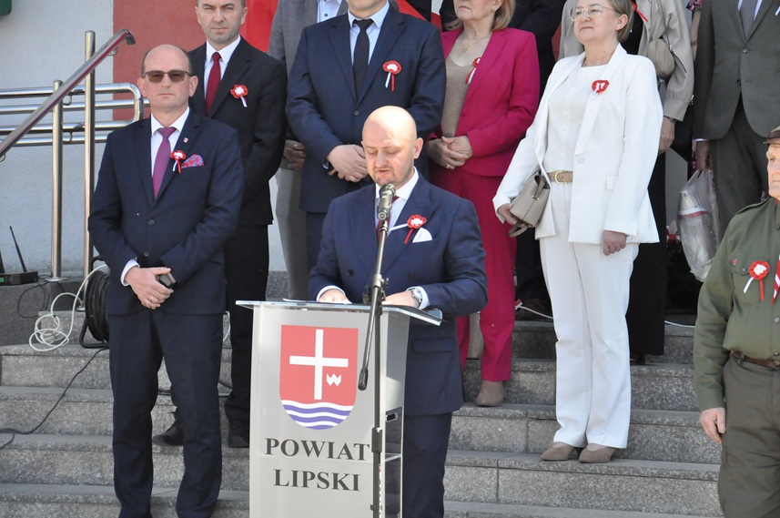 Mężczyzna w grafitowym garniturze stoi przy mównicy z herbem powiatu. Za nim na schodach stoi więcej osób w odświętnych ubraniach.