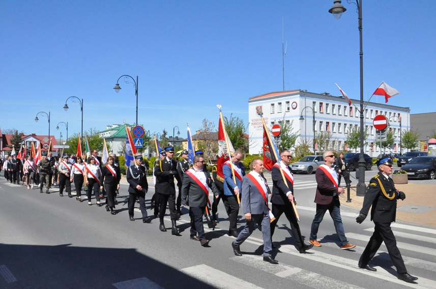 Duża grupa osób ze sztandarami w odświętnych strojach, niektórzy mają mundury strażackie, maszeruje ulicami miasta. pogodny dzień.