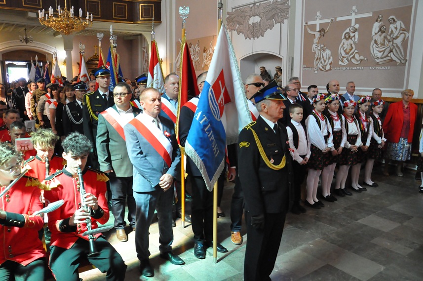Duża grupa ludzi. Orkiestra dęta, osoby ze sztandarami i ludzie w odświętnych strojach we wnętrzu kościoła.