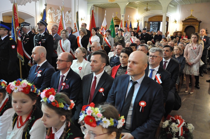 Duża grupa w odświętnych strojach siedzi na ławkach w kościele. obok nich stoją osoby ze sztandarami.