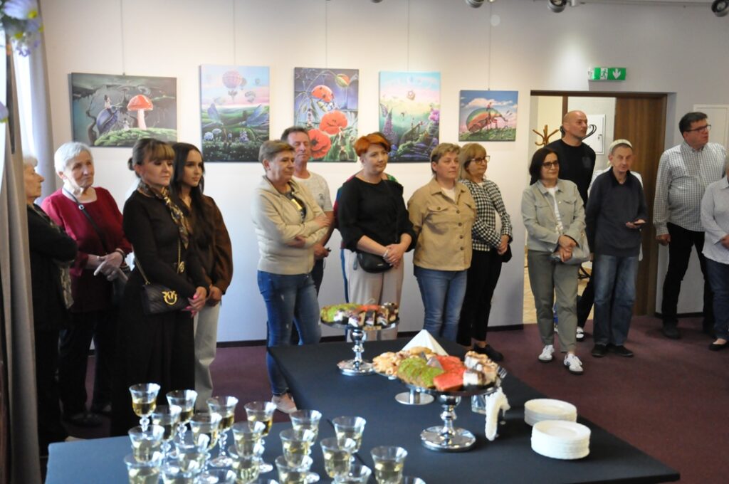 Grupa mężczyzn i kobiet stoi obok stołu z ciastami i kieliszkami. Za nimi kolorowe obrazy. Pomieszczenie ciepło oświetlone.