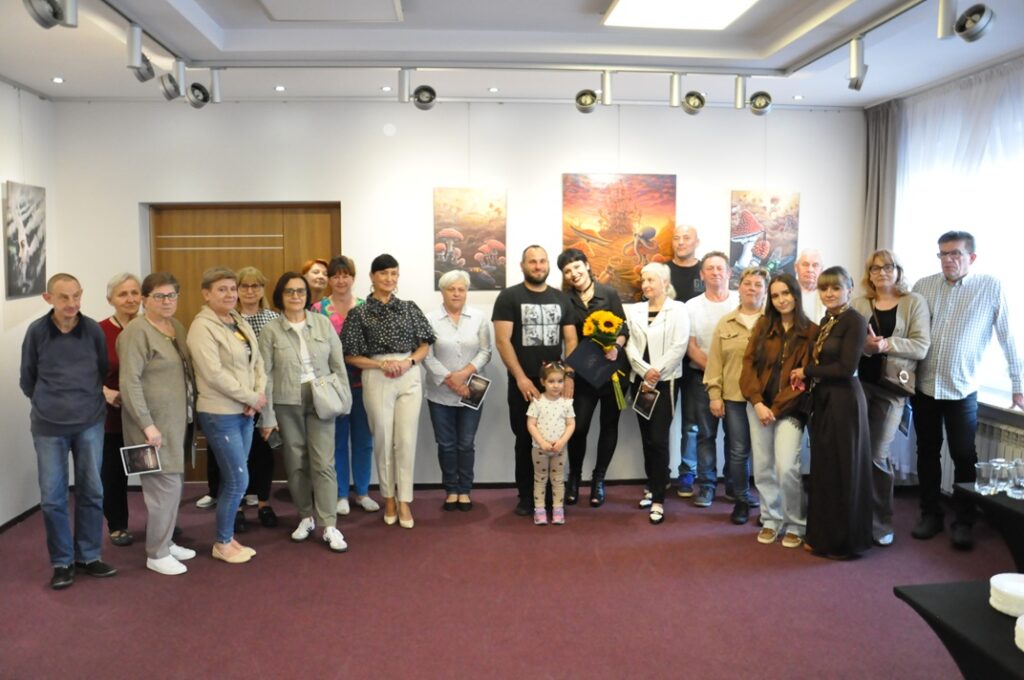 Duża grupa osób, dorośli i dziewczynka pozują do wspólnego zdjęcia. Jedna kobieta trzyma bukiet słoneczników. Niektórzy trzymają kolorowe kartki. Za nimi kilka baśniowych obrazów na białej ścianie i drzwi. Pomieszczenie ciepło oświetlone.