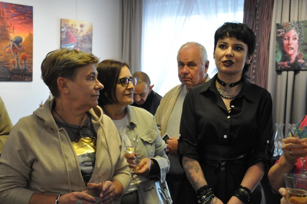 Kobieta w czarnym ubraniu z czarnymi bransoletami na obu rękach i ciemnym makijażu stoi obok kilku dorosłych osób. W tle na ścianach wiszą kolorowe obrazy. Pomieszczenie ciepło oświetlone.