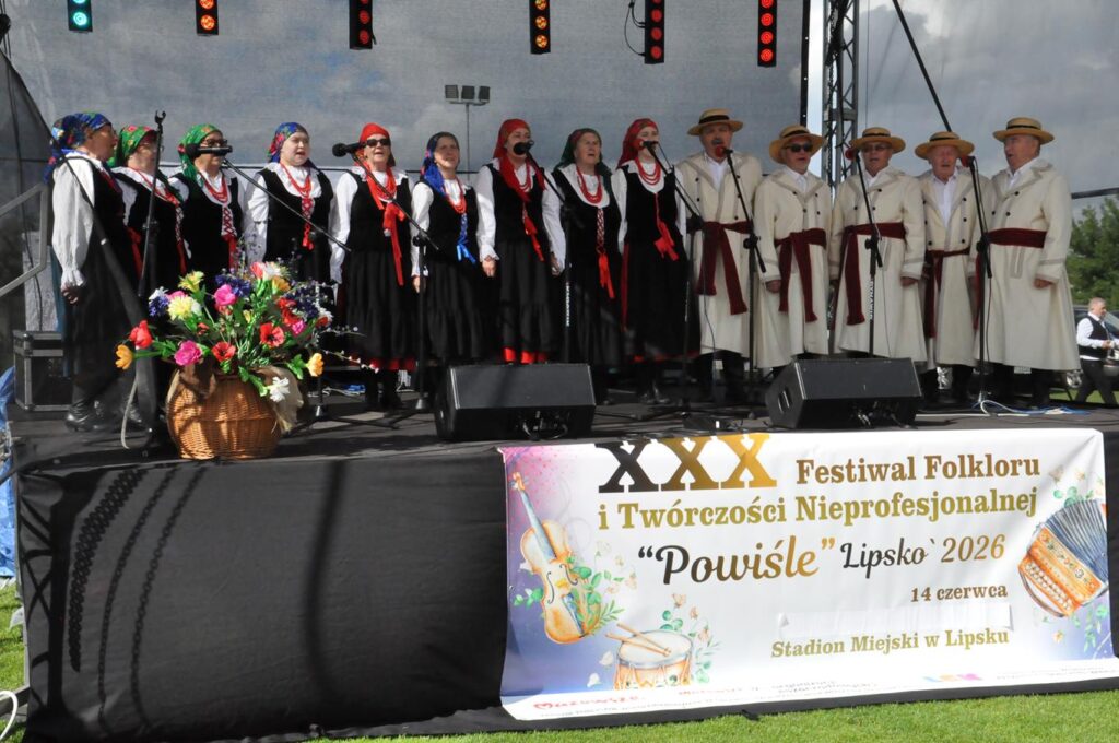 Duża grupa osób w ludowych strojach stoi na scenie przy mikrofonach na statywach. Na dole sceny wisi baner z nazwą wydarzenia. pogodny dzień.