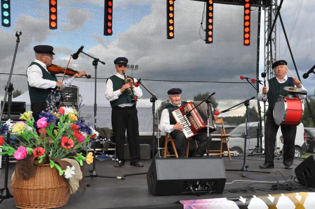 Grupa czterech ludowych muzyków z instrumentami stoi lub siedzi na scenie przy mikrofonach na statywach. Pochmurny dzień.