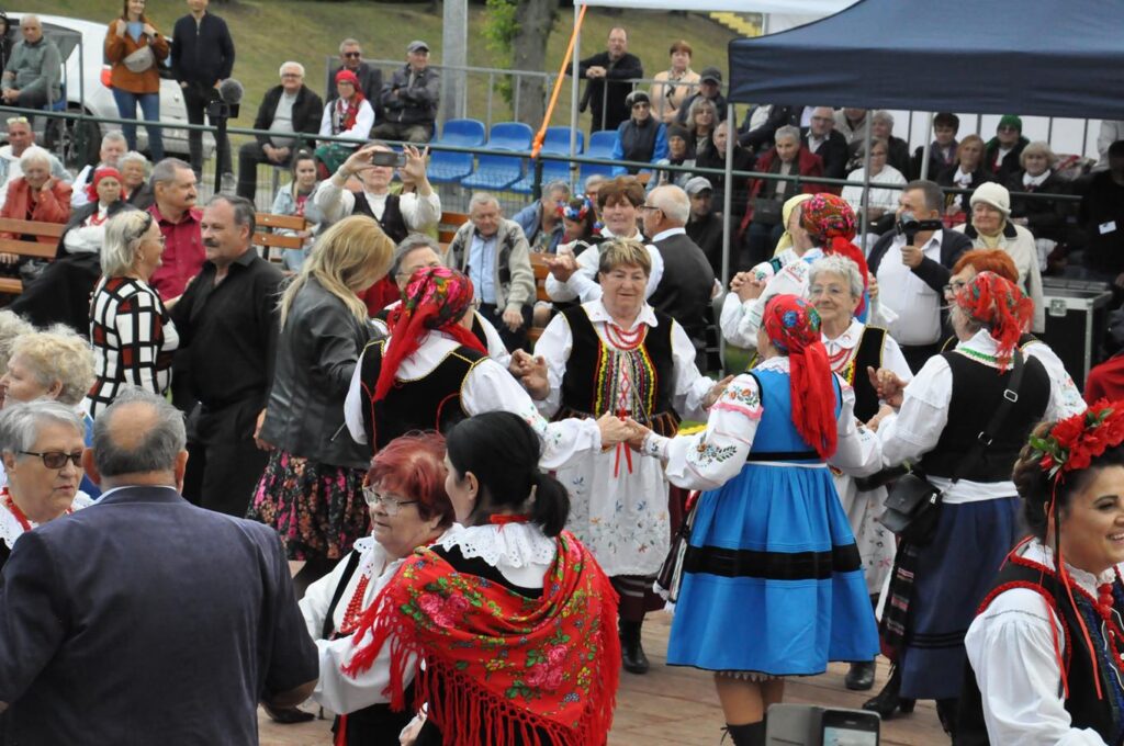 Duża grupa osób w ludowych strojach tańczy. Za nimi siedzą ludzie. Pochmurny dzień.