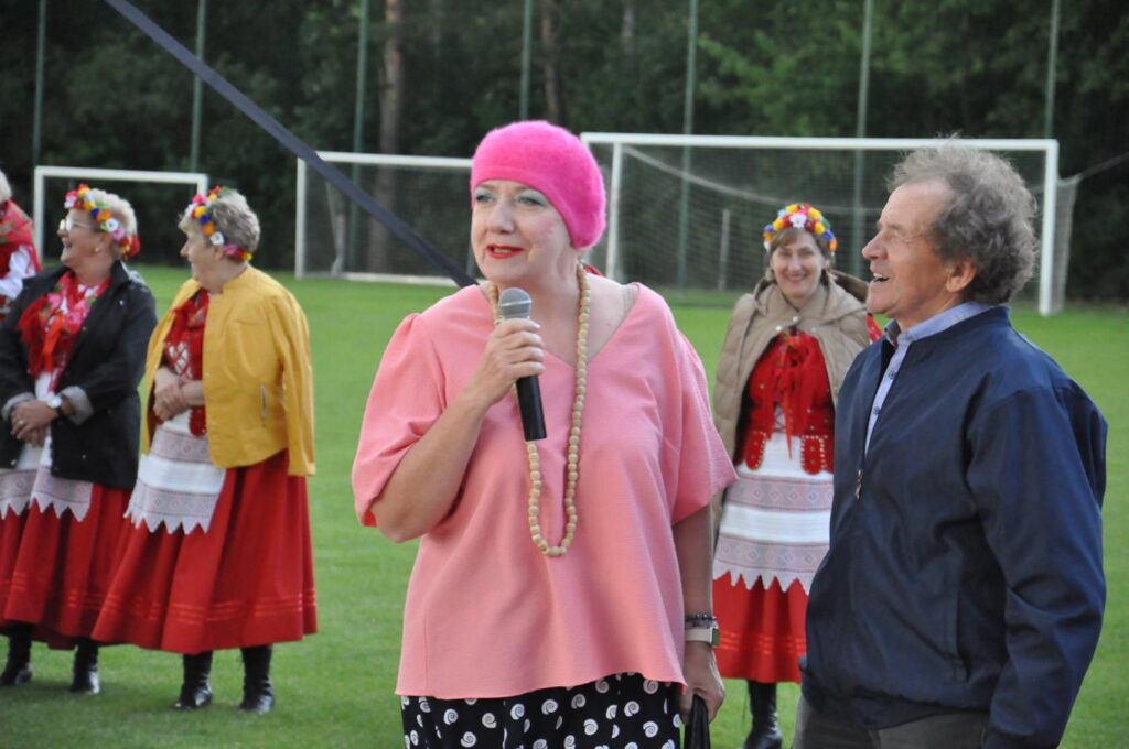 Kobieta w różowej koszulce i różowym berecie trzyma mikrofon przy ustach obok niej stoi mężczyzn w granatowej bluzie. Za nimi kilka osób w ludowych strojach. Pochmurny dzień.
