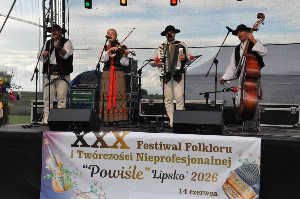 Grupa muzyków w ludowych strojach -kobieta i trzech meżczyzn stoi na scenie przy mikrofonach na statywach. Wszyscy trzymają instrumenty. Na dole sceny baner z nazwą wydarzenia. Pochmurny dzień.