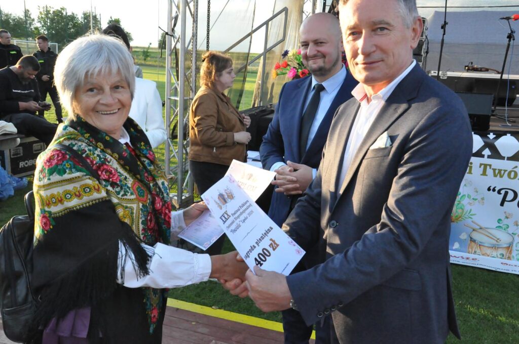 Mężczyzna w grafitowym garniturze podaje dłoń kobiecie z chustą na ramionach. W drugiej dłoni trzyma kartkę z kwotą nagrody pieniężnej. Za nimi stoi kilka osób przed sceną.