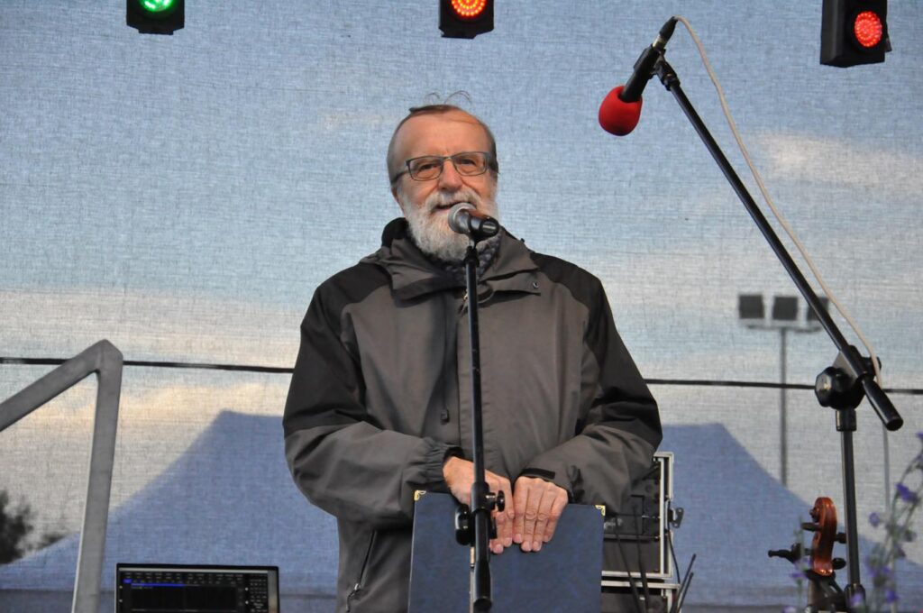 Mężczyzna w szarej kurtce z brodą i w okularach stoi na scenie przy mikrofonie na statywie.