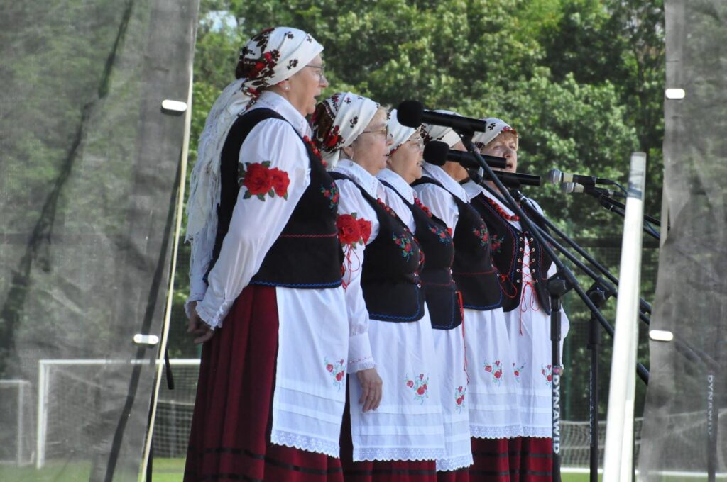 Kilka kobiet w ludowych strojach śpiewa przy mikrofonach na statywach. W tle drzewa.