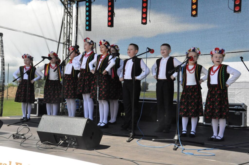 Duża grupa dzieci w ludowych strojach na scenie śpiewa do mikrofonów na statywach. Pogodny dzień.
