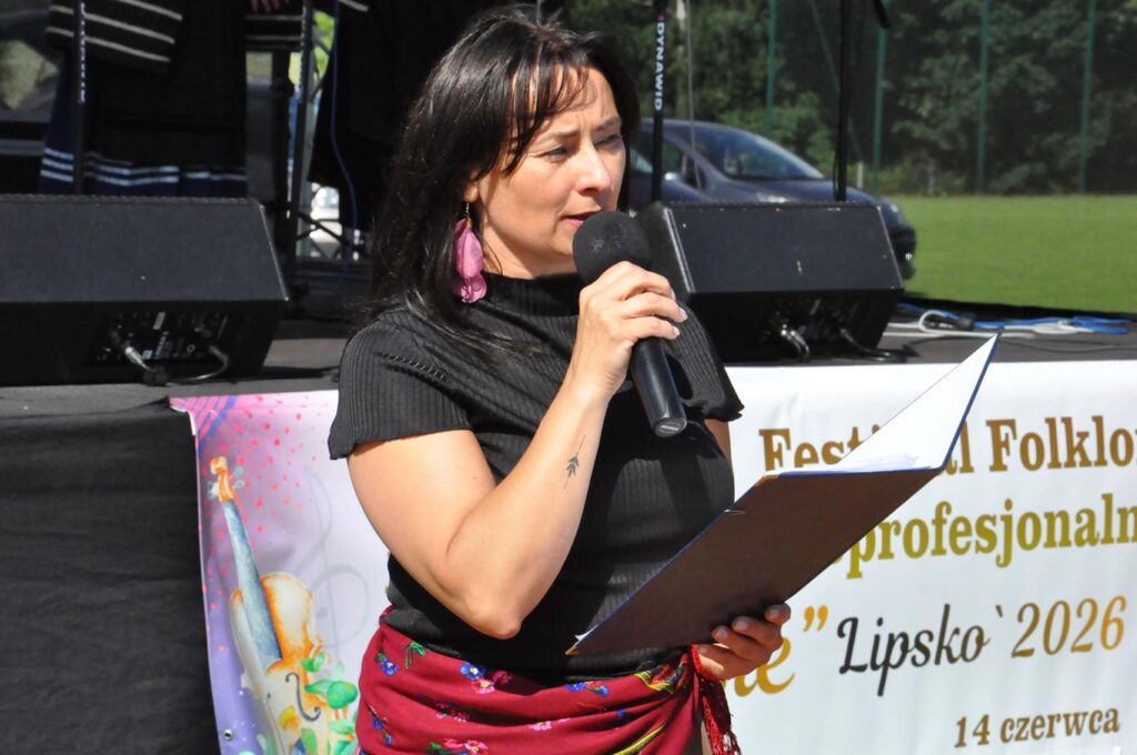 Kobieta w czarnej koszulce w jednej ręce trzyma mikrofon przy ustach, a w drugiej podkładkę konferansjerską. Za nią scena. pogodny dzień.