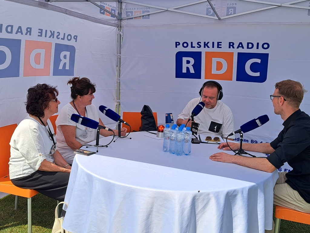Cztery dorosłe osoby siedzą przy mikrofonach na statywach na okrągłym stole przykrytym białym obrusem. Na stole są też butelki wody mineralnej. W tle logo rozgłośni radiowej na białych ścianach namiotu.