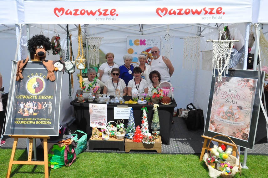 Dwa plakaty w ramkach na stelażach wystawienniczych po obu stronach. W środku grupa osób pozuje do zdjęcia przy stole z ekspozyjdą z rękodziełem. Nad nimi logotypy organizatora.