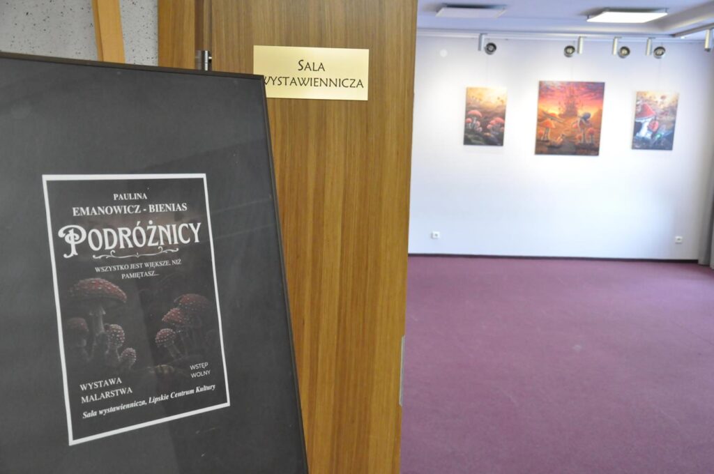 Stelaż z plakatem promującym wystawę. W dalszym planie drzwi wejściowe do sali wystawienniczej oraz ściana z trzema obrazami.