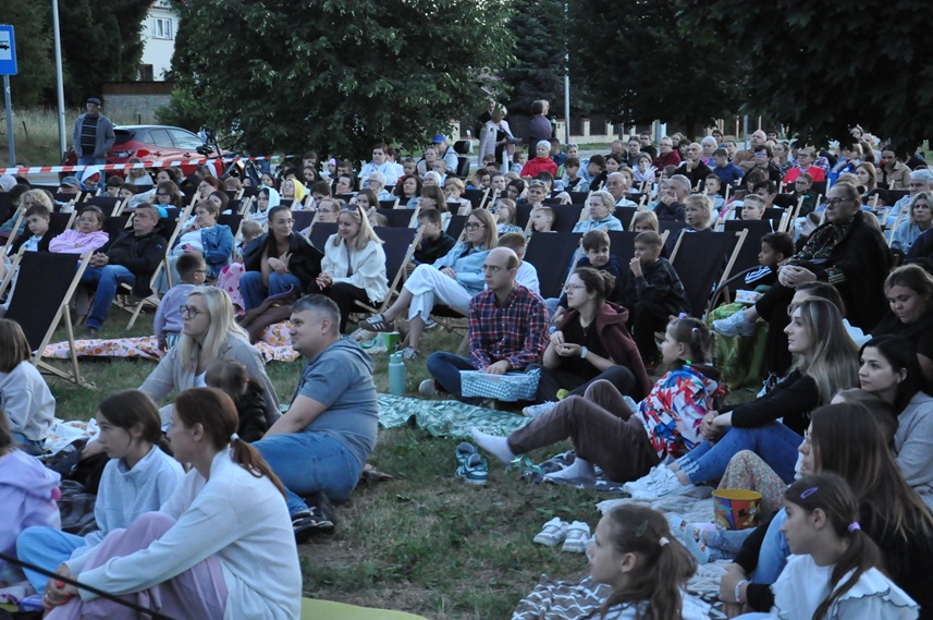 Duża grupa ludzi siedzi na krzesłach i leżakach. Niektórzy też na kocach. Za nimi drzewa. Plener wieczorowy. Pogodny dzień.