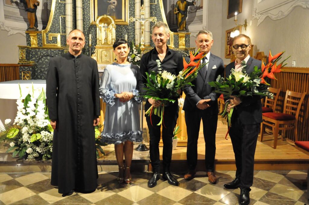 Ksiądz kobieta w błękitnej sukience i trzech mężczyzn w galowych strojach pozują do wspólnego zdjęcia. Dwóch mężczyzn trzyma kwiaty. Wnętrze kościoła.