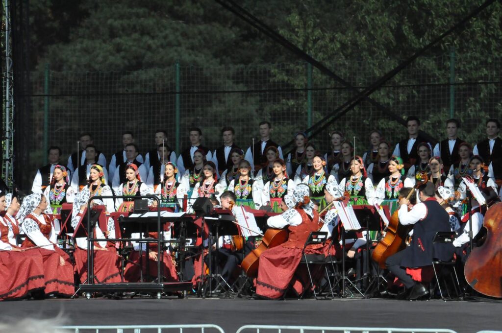 Duża grupa osób w ludowych strojach na scenie plenerowej. Większość stoi w kilku rzędach. przed nimi pozostałe osoby siedzą i trzymają instrumenty muzyczne.