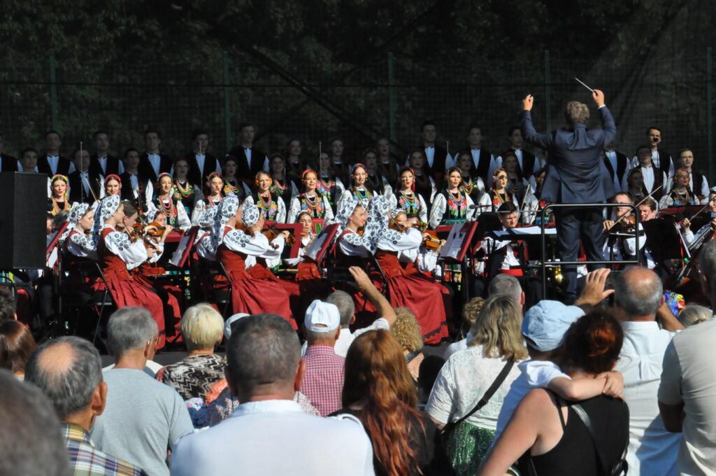 Dyrygent w czarnym garniturze wśród muzyków w strojach ludowych. Za nim publiczność. Pogodny dzień.