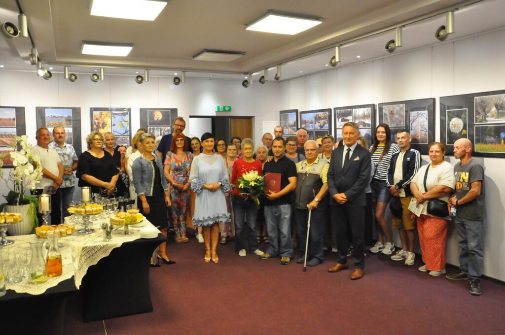 Duża grupa osób pozuje do wspólnego zdjęcia. Wszyscy stoją. Za nimi antyramy z kolorowymi fotografiami. Pomieszczenie ciepło oświetlone.